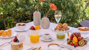 Une table dressée avec des aliments pour le petit-déjeuner, notamment des crêpes, des croissants et des fruits, et offrant une vue panoramique sur un magnifique paysage viticole.