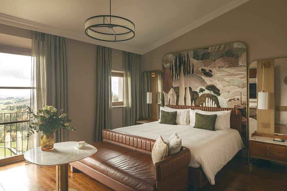 Ett hotellrum med en stor säng, en sänggavel i trä, ett runt bord med en vas med blommor och utsikt över landskapet.