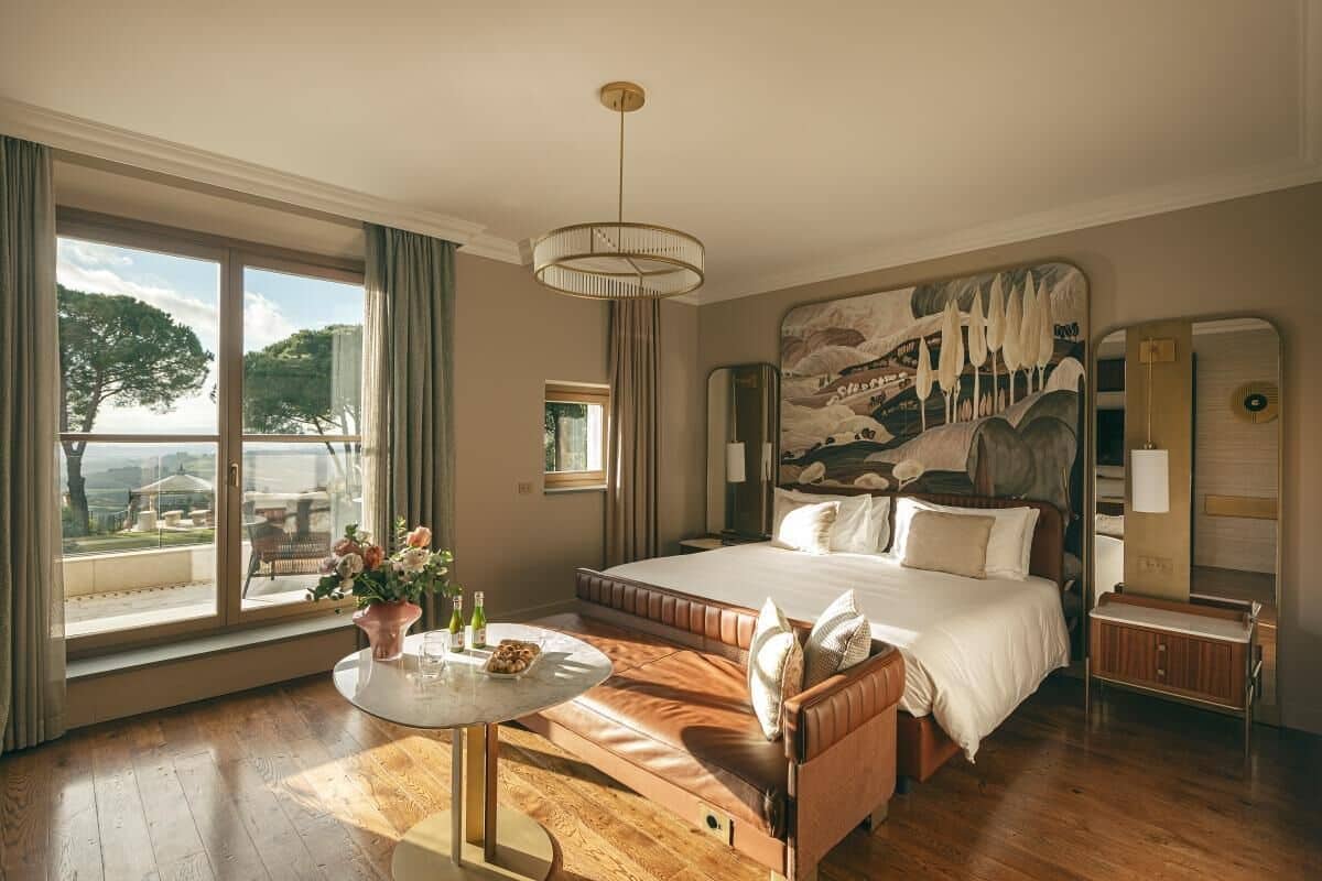 Ett rymligt sovrum med en stor säng, ett runt bord med en vas med blommor och utsikt över naturen genom ett stort fönster.