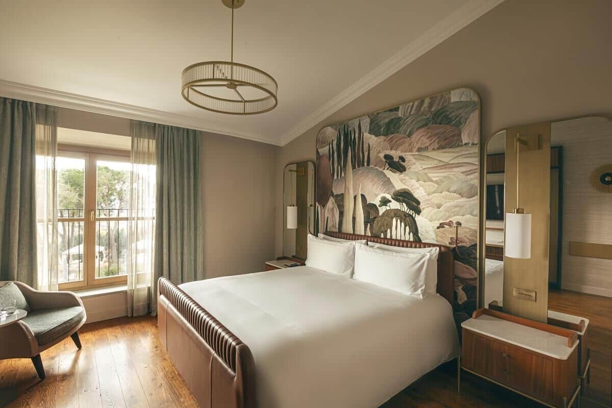 Ett hotellrum med en stor säng, en sittgrupp med en fåtölj, ett stort fönster med gardiner, en tavla på väggen och en spegel.