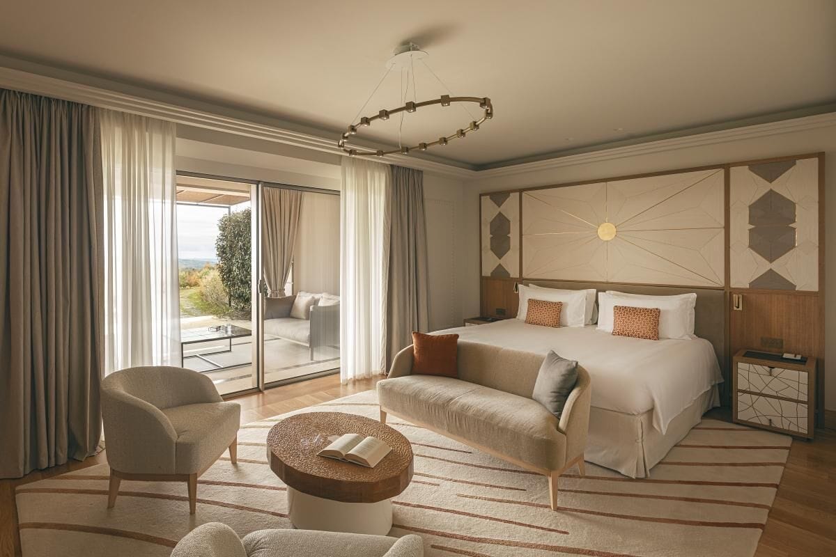 Ett modernt hotellrum med en stor säng, en soffa, en fåtölj och ett litet bord. Genom glasdörren har man utsikt över omgivningarna.