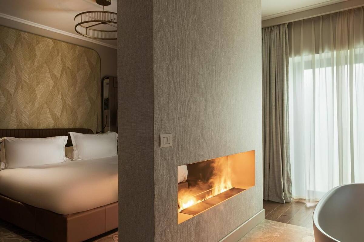 Ett modernt hotellrum med en stor säng, en inbyggd öppen spis och utsikt över omgivningarna genom tunna, genomskinliga gardiner.
