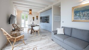A modern living room with a grey sofa, wicker chairs, a dining table with chairs, a kitchen area, and a mounted TV.