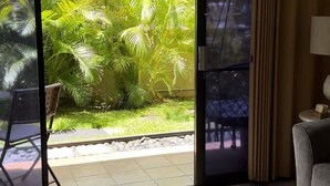A room with a sliding glass door leading to a garden with palm trees. There is a round table with decorative items and a chair. - 2 Bedroom Ocean Jeep  (Kihei)