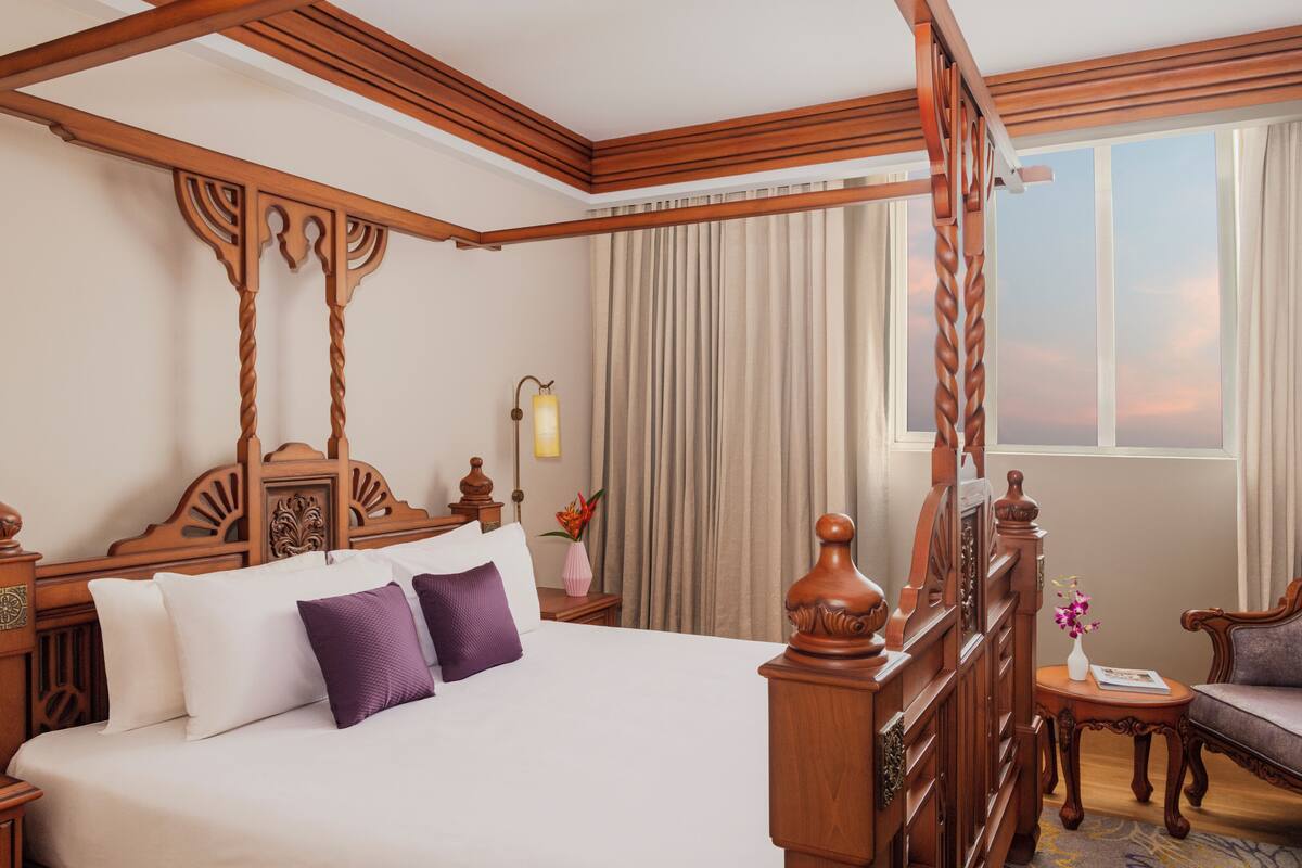 Una cama con dosel con almohadones blancos y morados, una mesita de noche de madera con una lámpara, y vista al atardecer a través de un ventanal grande.