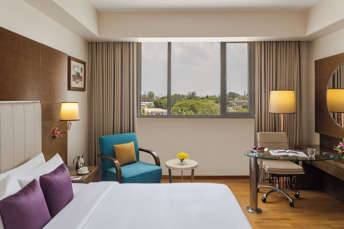 Habitación de hotel con una cama grande, un escritorio con silla, una mesita redonda con un jarrón de flores y vistas a la ciudad a través de amplias ventanas.