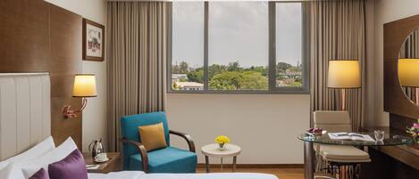 A hotel room with a large bed, a desk with a chair, a small round table with a vase of flowers, and a view of the city through large windows.