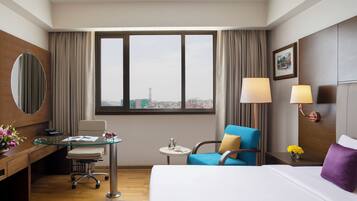 A hotel room with a bed, a desk with a chair, a small table with a lamp, and a view of the city through a large window.