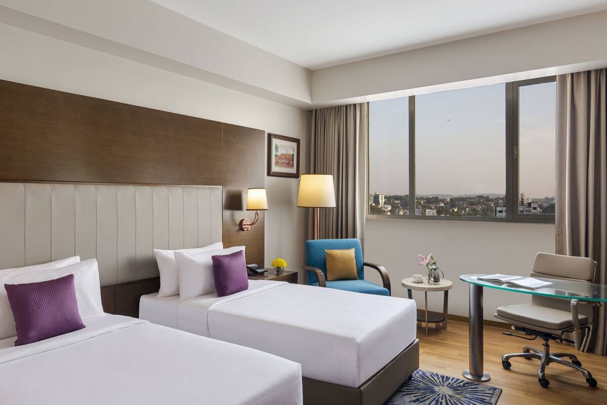 Habitación de hotel con dos camas, un escritorio, una silla, una lámpara, una ventana con vistas a la ciudad y una mesita con un florero.