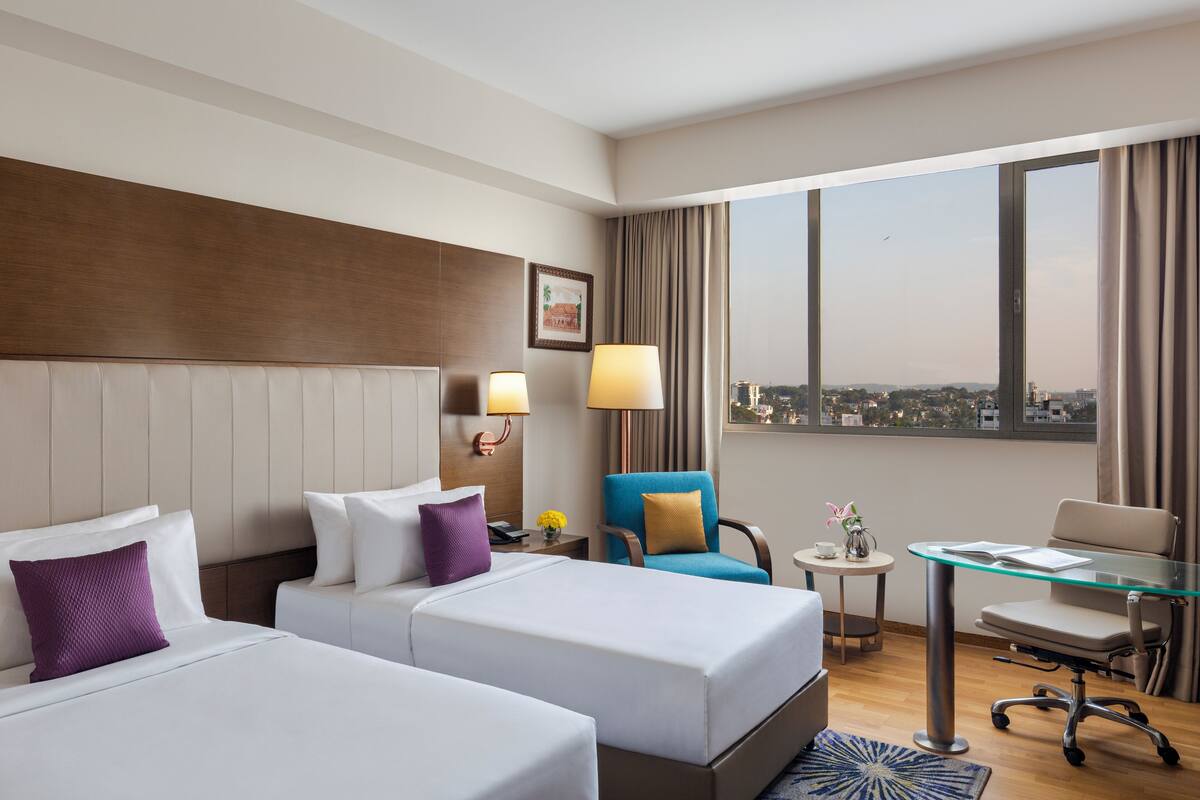 Habitación de hotel con dos camas, un escritorio, una silla, una mesita de noche con lámpara y vistas a la ciudad.