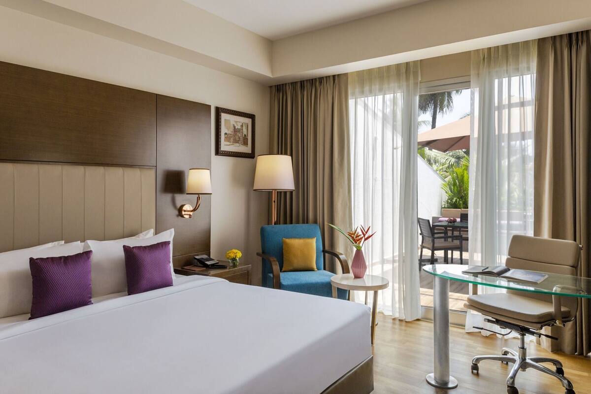 Habitación de hotel con una cama grande, dos lámparas de noche, una silla, una mesita y un balcón con vistas.