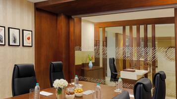 Un salón de conferencias con una mesa de madera, sillas negras, un jarrón con flores y botellas de agua.