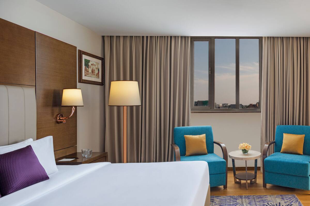 Una habitación de hotel con una cama, dos sillones, una mesita con un jarrón y una ventana con cortinas.