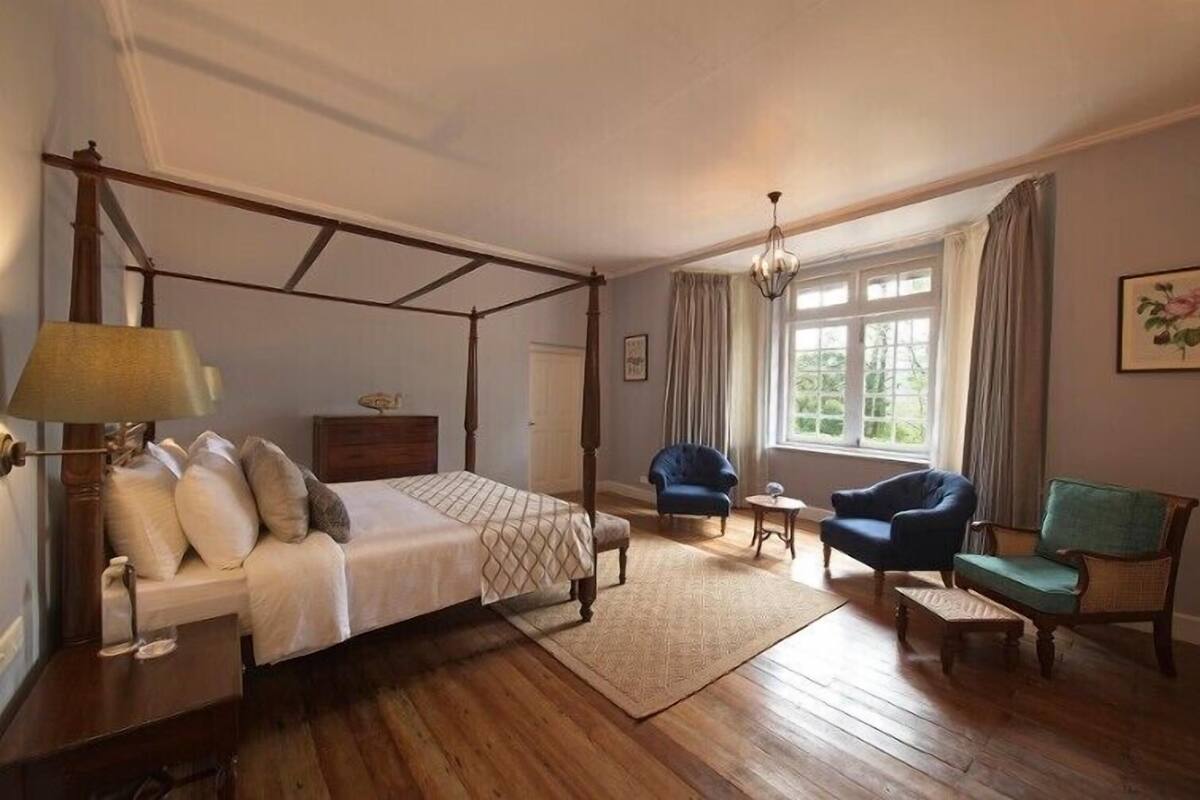 A four-poster bed with a wooden frame, a bedside table with a lamp, a small round table, two upholstered chairs, and a window with curtains.