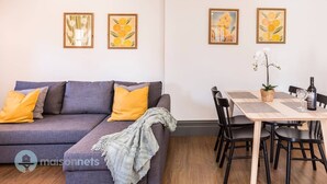 Ein modernes Wohnzimmer mit einem grauen Sofa, gelben Kissen und einem Essbereich mit Tisch, Stühlen und gerahmten Bildern an der Wand.