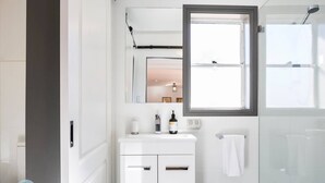 Ein modernes Badezimmer mit einem weißen Waschtisch, einem großen Spiegel, einem Fenster und einer Glasdusche.