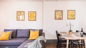 A modern living room with a grey sofa, yellow cushions, and a dining area with a table, chairs, and framed artwork on the wall.