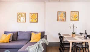 A modern living room with a grey sofa, yellow cushions, and a dining area with a table, chairs, and framed artwork on the wall.
