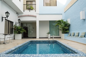 Una piscina con bordi piastrellati, circondata da un edificio bianco e blu con un balcone, piante in vaso e un'area salotto con cuscini.