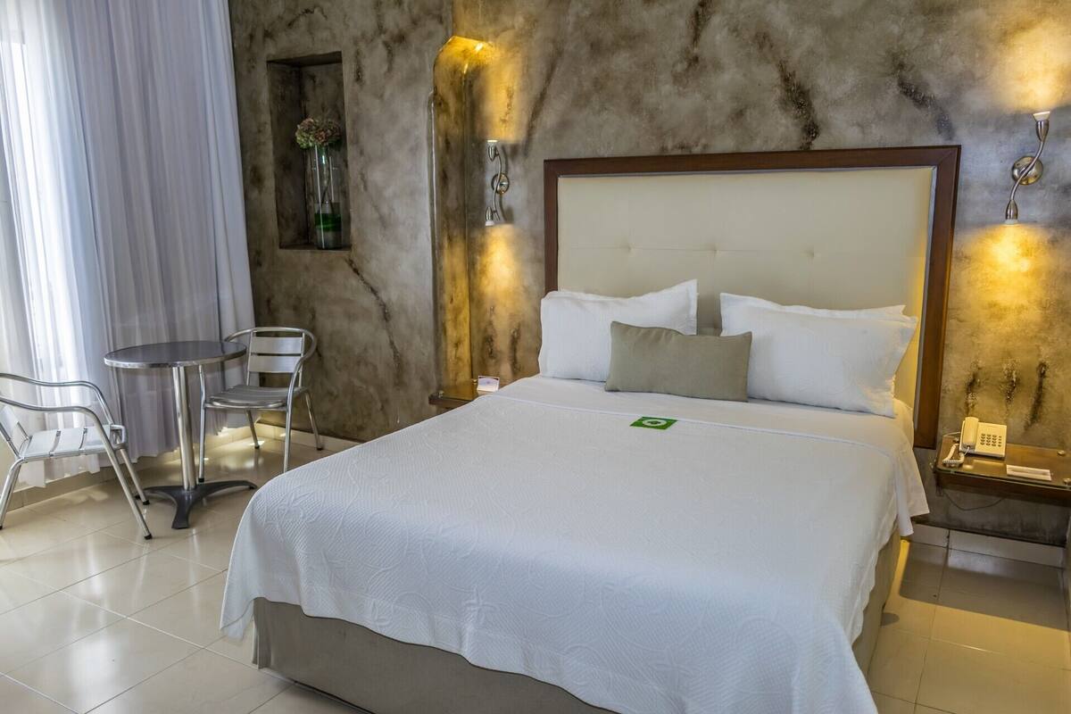 A hotel room with a bed, a chair, a small table, and a wall-mounted lamp.