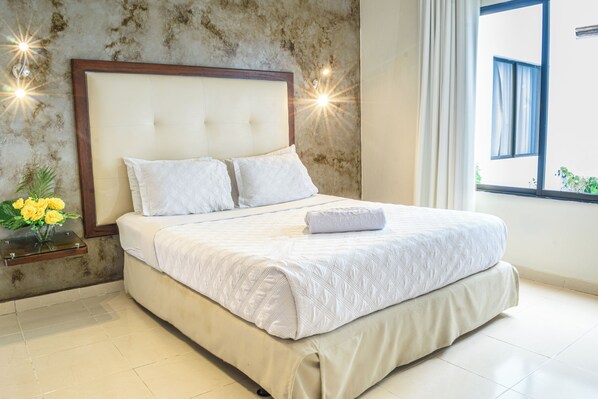 Una cama bien hecha con edredón blanco y almohadas, una mesita de noche con un jarrón de flores amarillas, y una ventana con cortinas.
