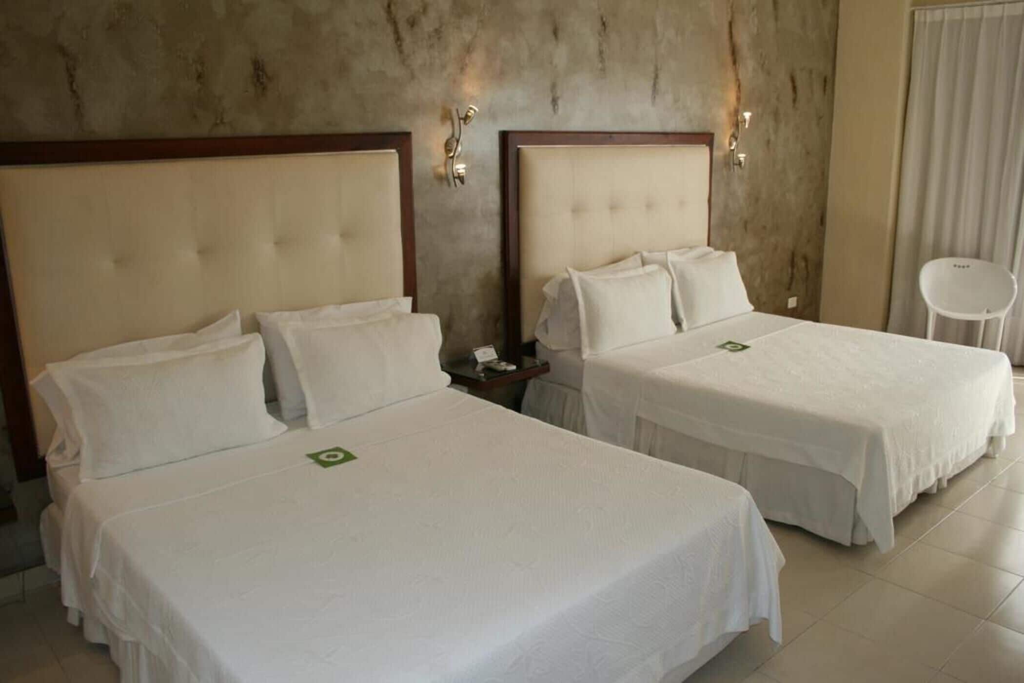 Habitación de hotel con dos camas, cada una con cabecero de madera y ropa de cama blanca. Hay luces fijadas a la pared y una pequeña mesa entre las camas.