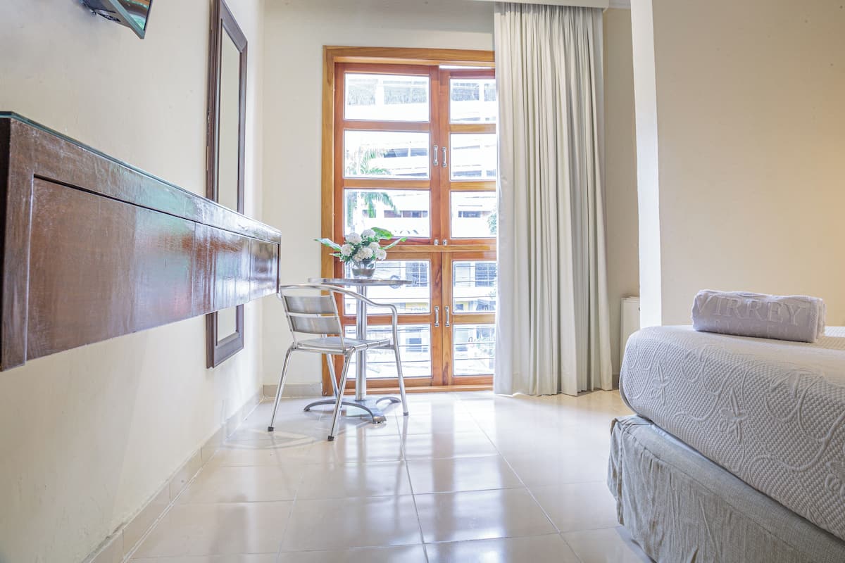 A hotel room with a bed, a chair, a table with a vase of flowers, and a large window with curtains.