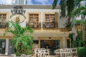 A hotel with a sign reading 'HOTEL VIRREY CARIAGEN,' featuring a balcony with wooden railings, a large circular emblem with a checkmark, and outdoor seating with white chairs. - Hotel Virrey Cartagena (Cartagena)