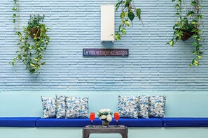 A blue brick wall with hanging plants, a sign with a Spanish phrase, a blue sofa with patterned cushions, and two red glasses on a small table. - Hotel Virrey Cartagena (Cartagena)