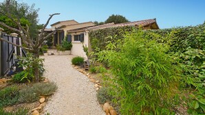 Un étroit sentier de gravier mène à une maison dont le toit est recouvert de tuiles, et qui est entourée d’une végétation luxuriante et d’une clôture.