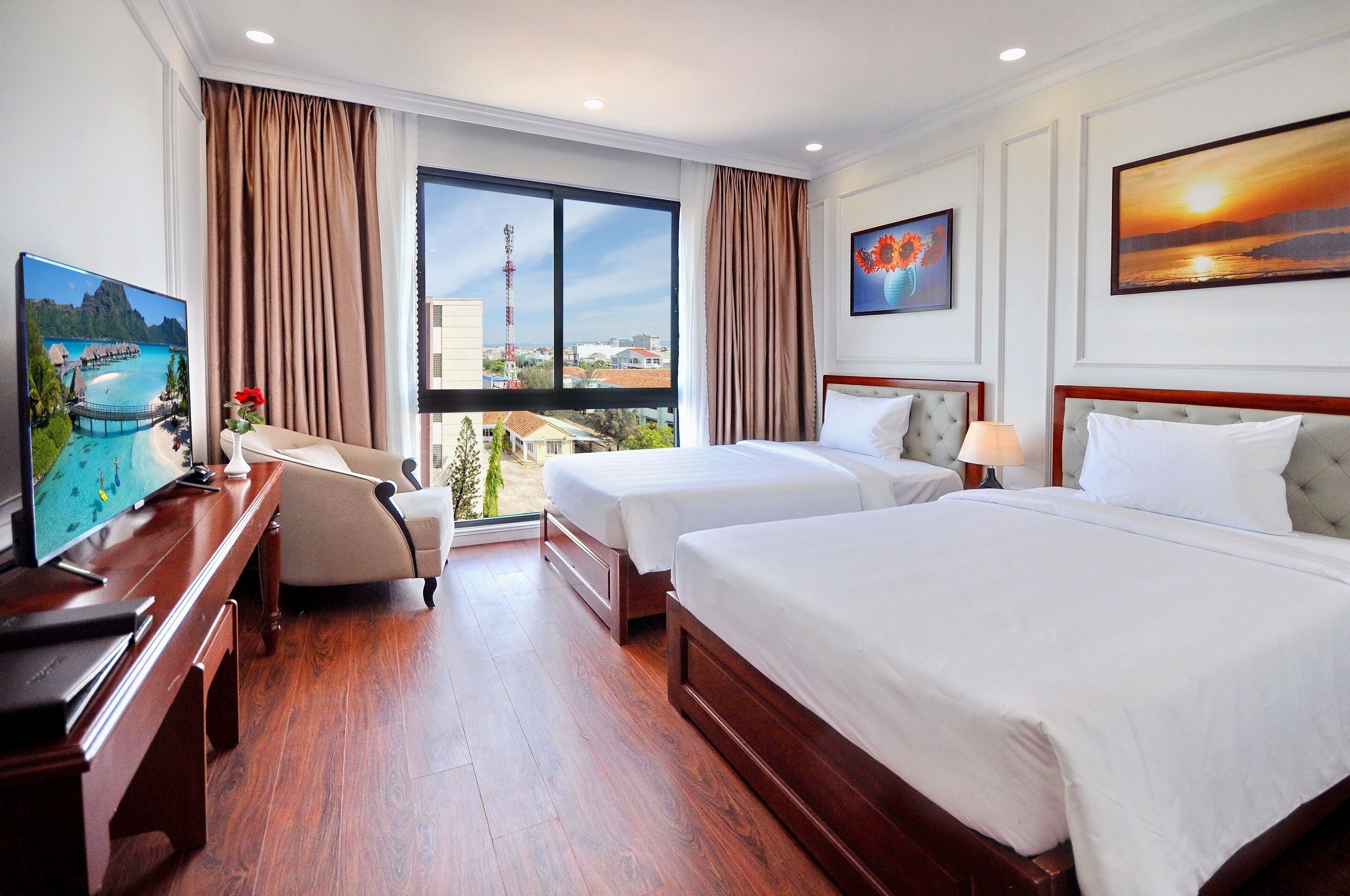 A hotel room with two beds, a desk with a TV, and large windows with curtains.