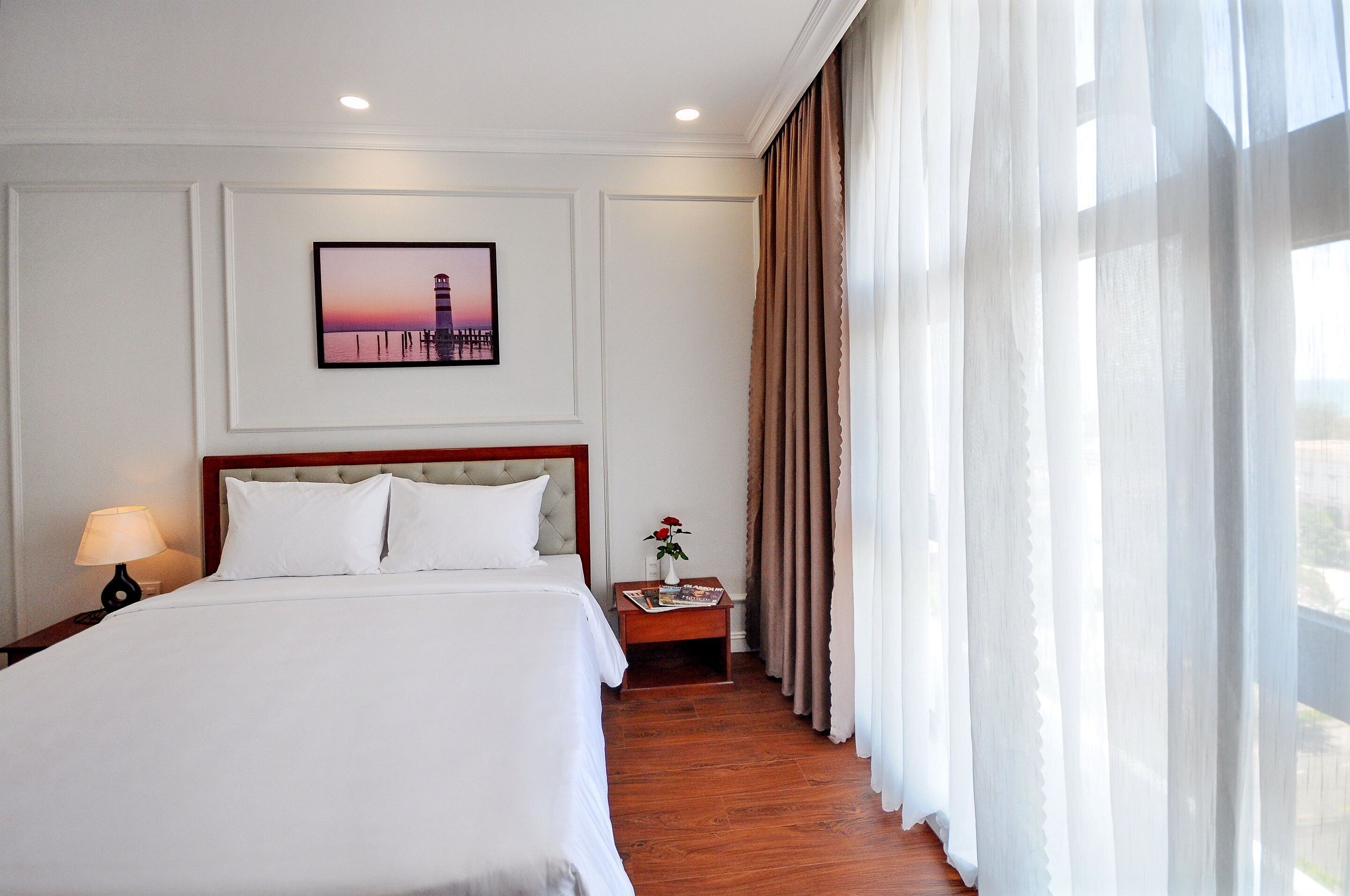 A hotel room with a large bed, a nightstand with a lamp, a framed picture on the wall, and a window with curtains.