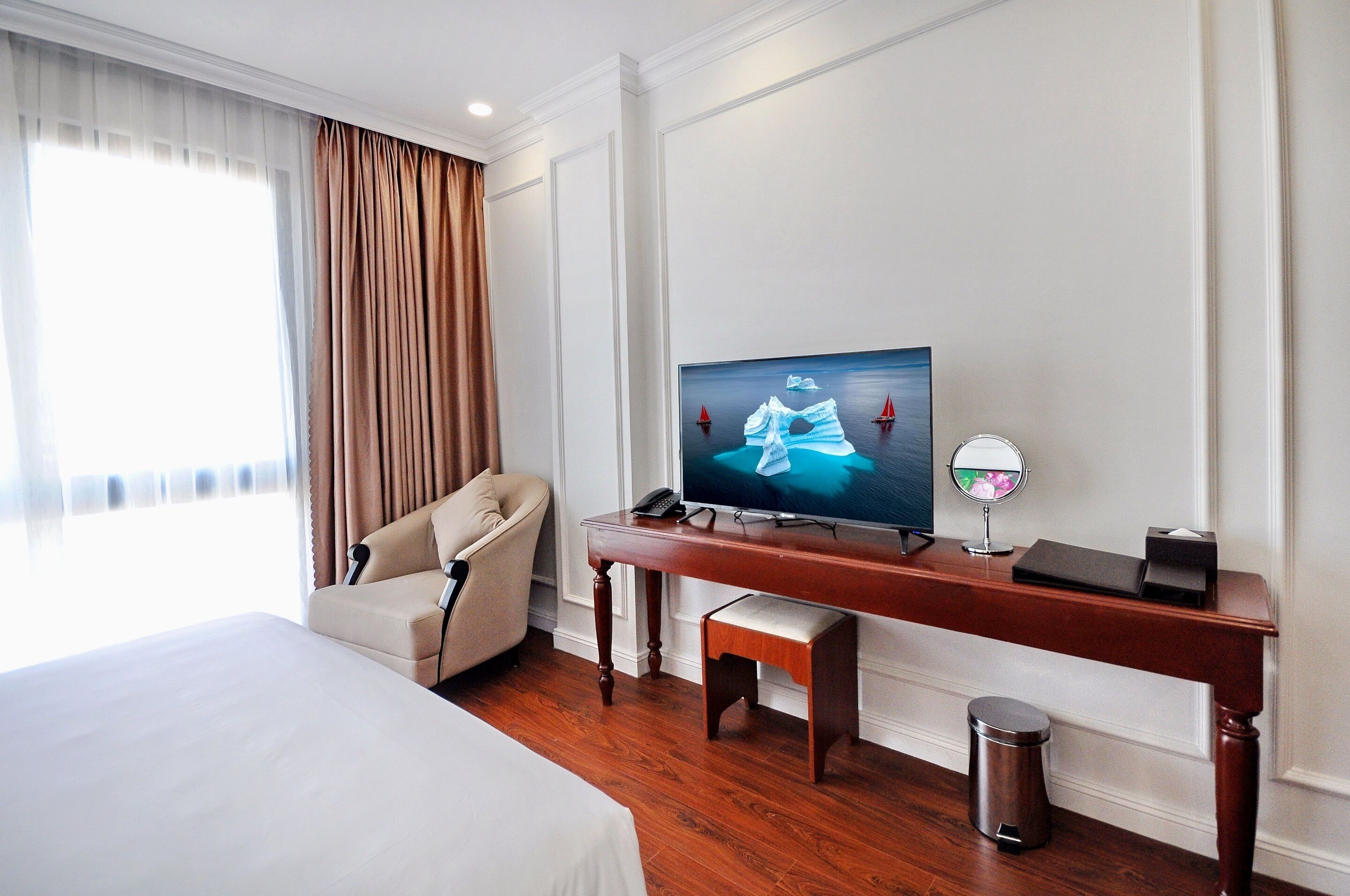 Ein Hotelzimmer mit Bett, Fernseher auf Holzfuß, Stuhl, kleinem Tisch und einem Fenster mit Vorhängen.