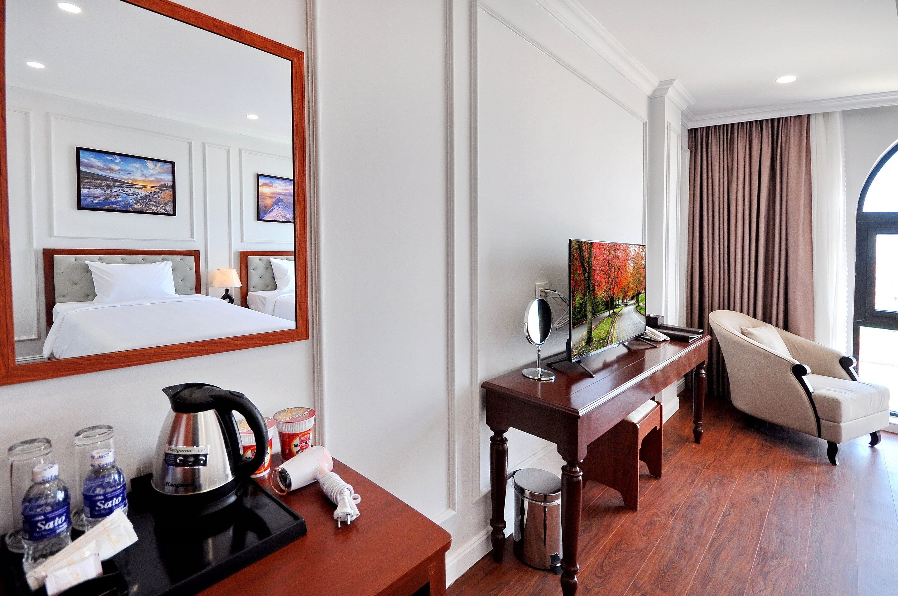 A hotel room with a bed, a desk with a kettle, a chair, and a large mirror.