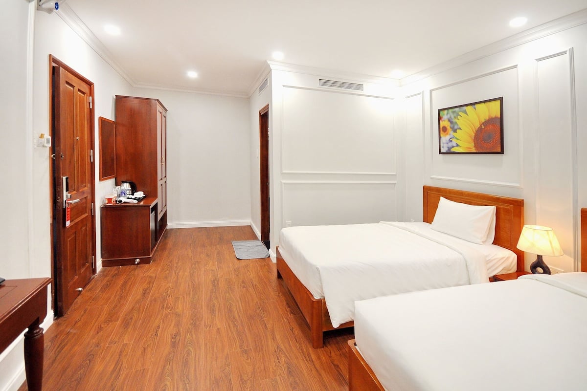 A hotel room with two beds, wooden flooring, a wardrobe, a desk, and a painting on the wall.