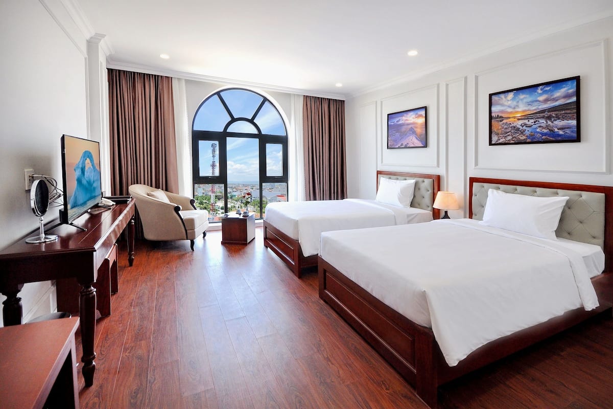 A hotel room with two beds, a desk with a computer, a chair, a large window with curtains, and artwork on the walls.
