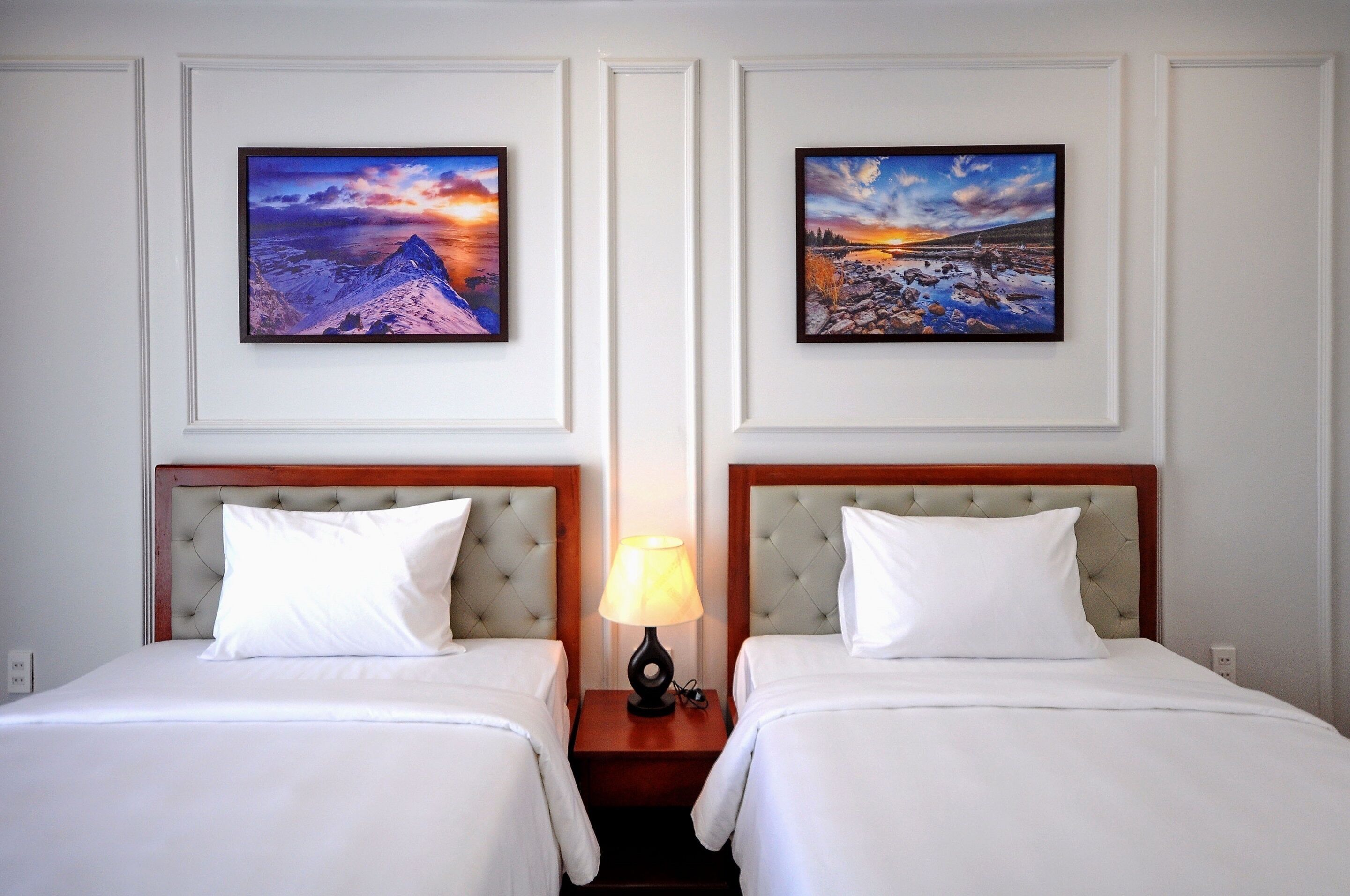 Une chambre d’hôtel avec deux lits, une table de chevet avec une lampe et deux tableaux représentant des paysages, encadrés et accrochés au mur.