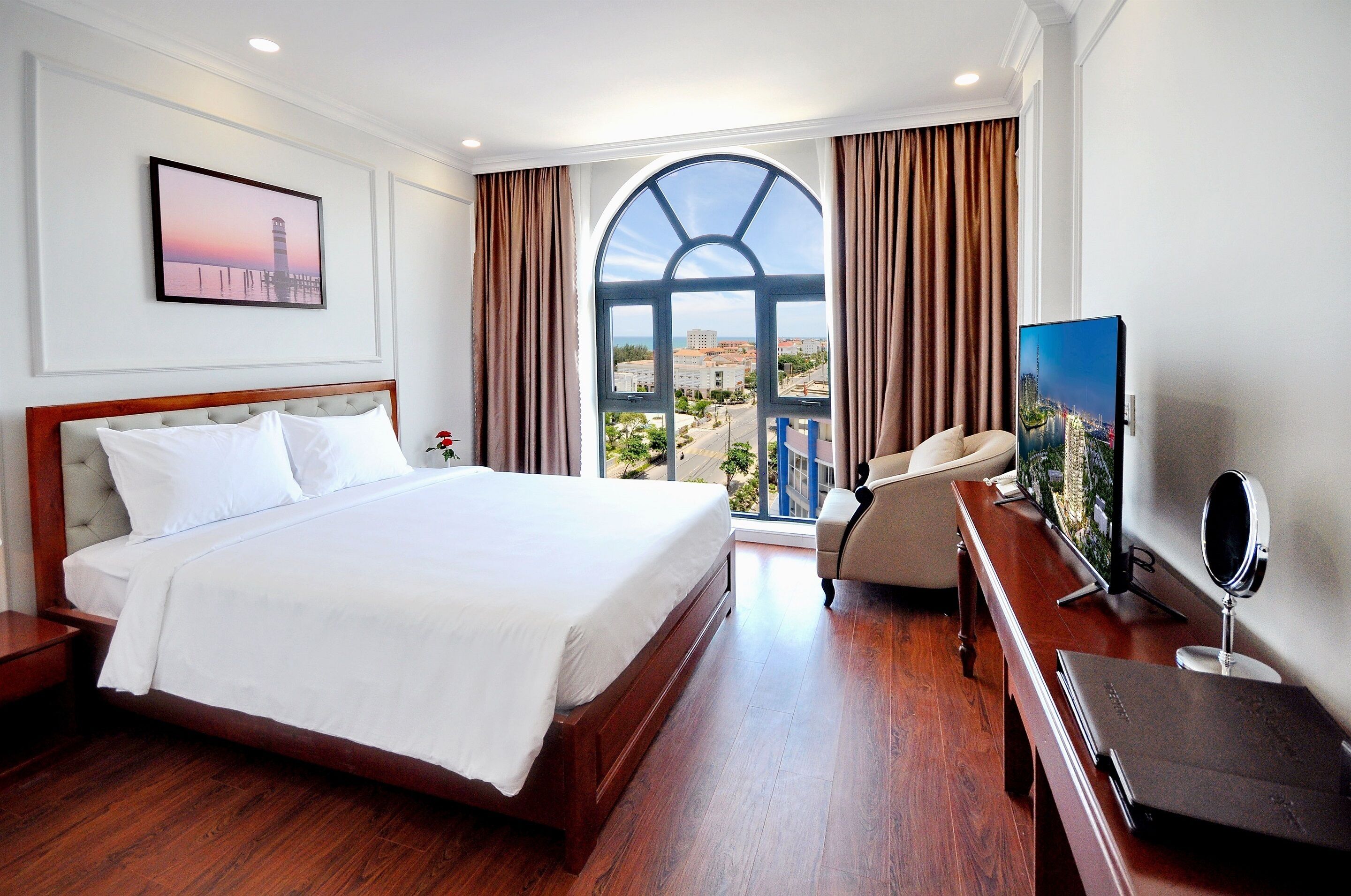Une chambre d’hôtel avec un grand lit, une télévision, un bureau avec un ordinateur et une vue sur le paysage urbain, visible à travers une grande fenêtre.