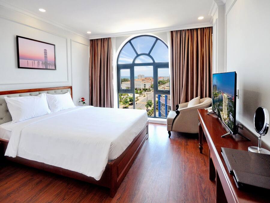 Quarto de hotel com uma cama grande, televisão, escrivaninha com computador e vista para a paisagem urbana através de uma janela ampla.