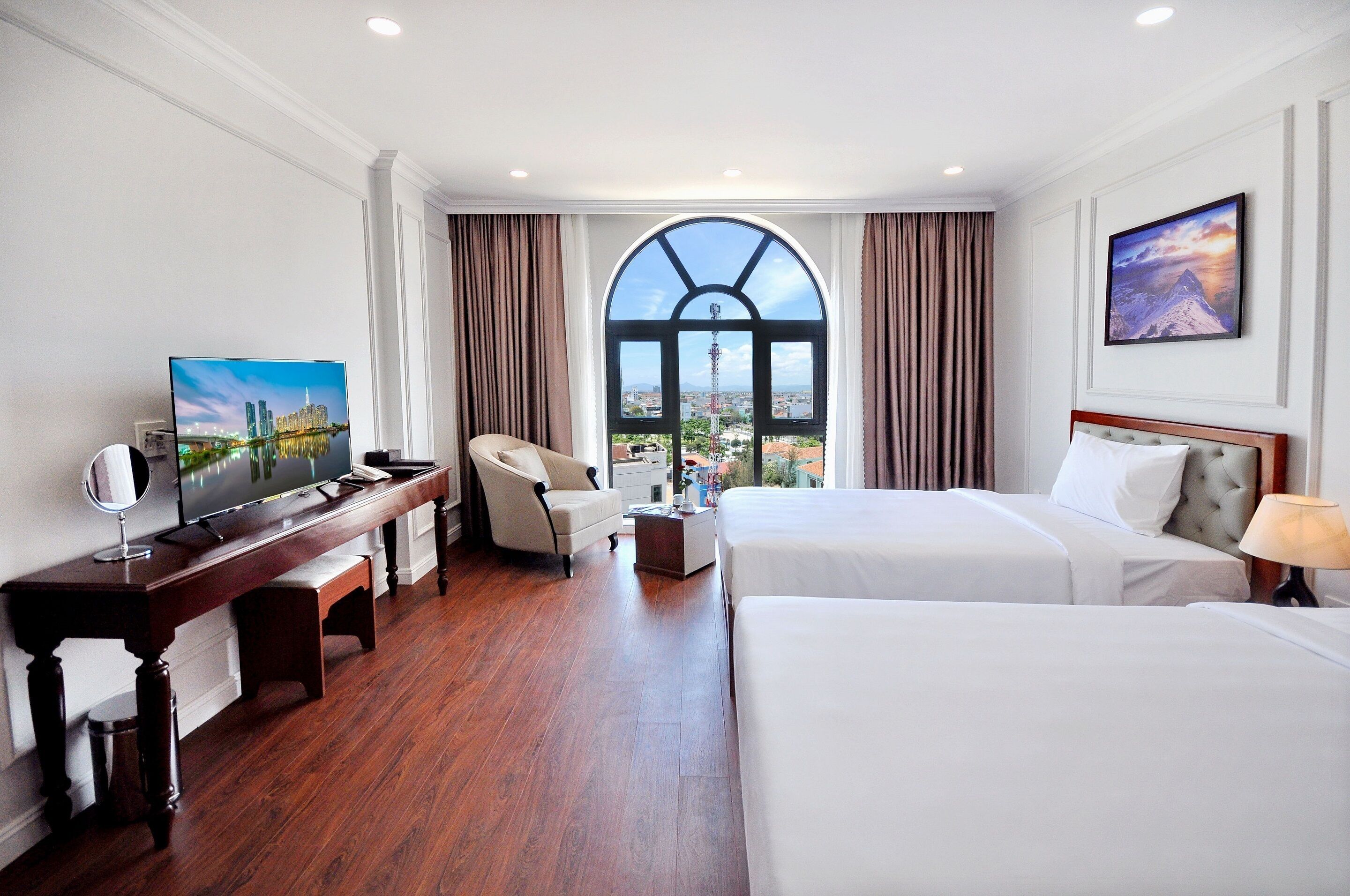 Habitación de hotel con un amplio ventanal de arco, cama, escritorio con televisor, silla y mesita de noche.