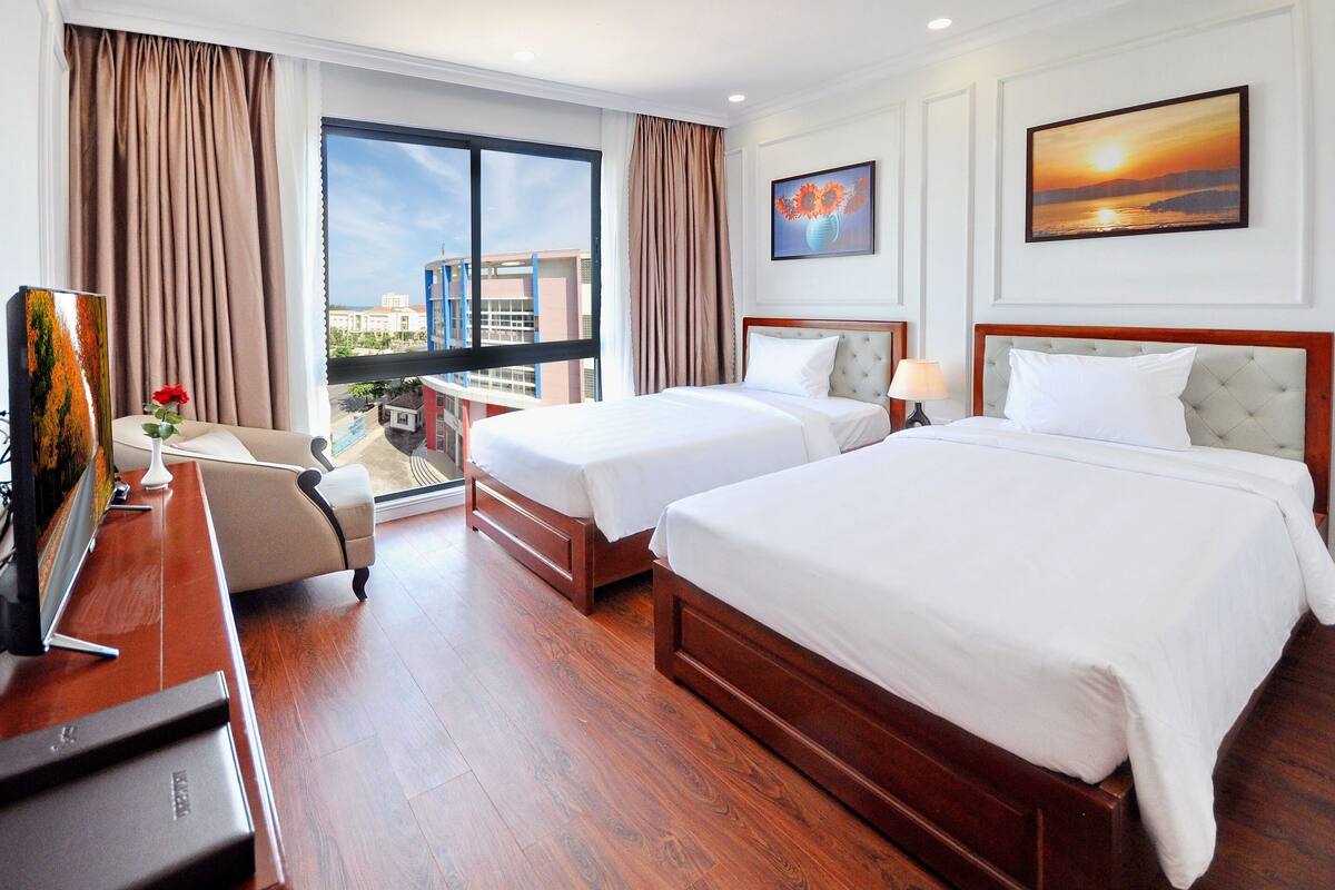 A hotel room with two beds, a desk, a chair, a TV, and a large window with a view of buildings.