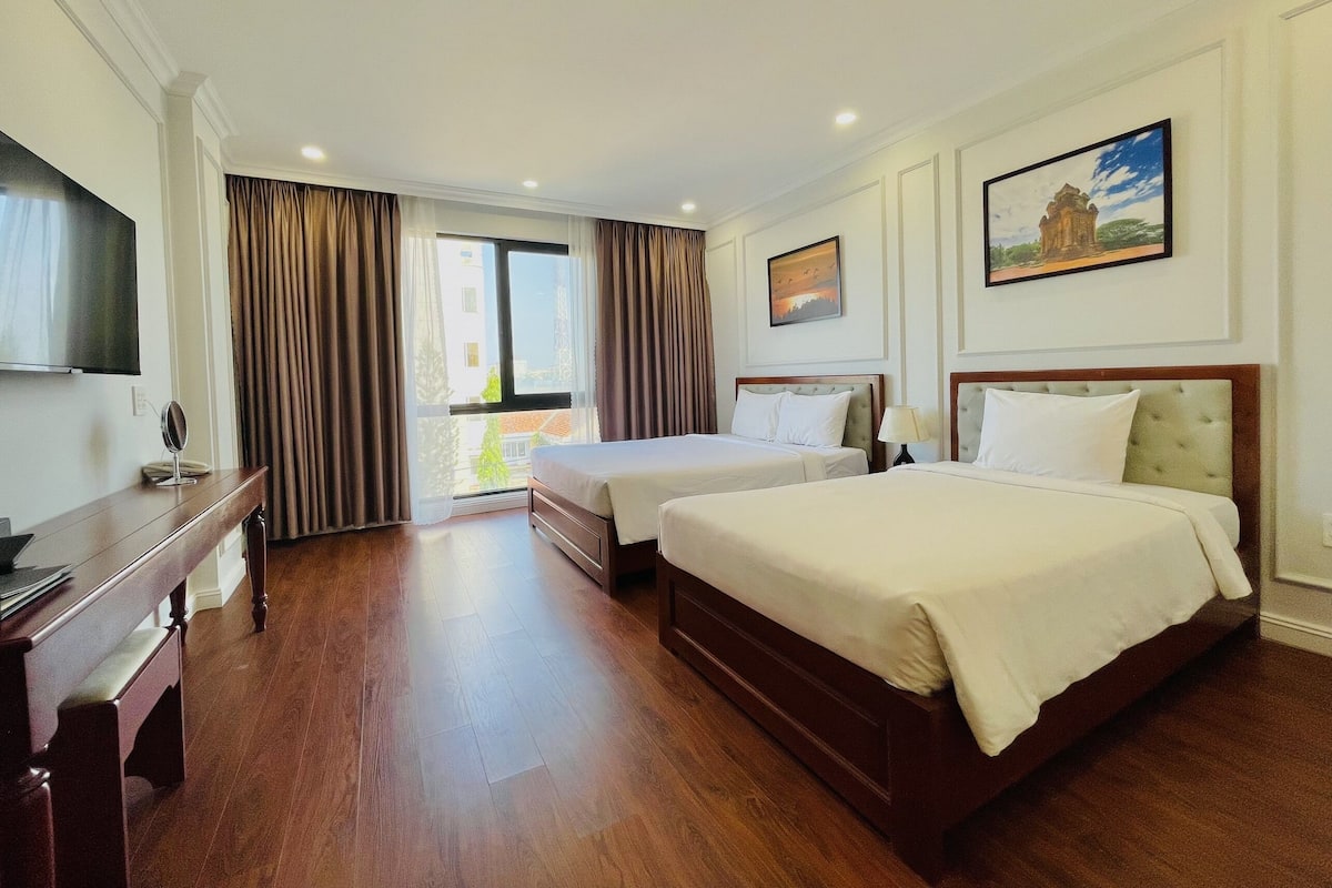A hotel room with two beds, a TV, a desk, and a window with curtains.