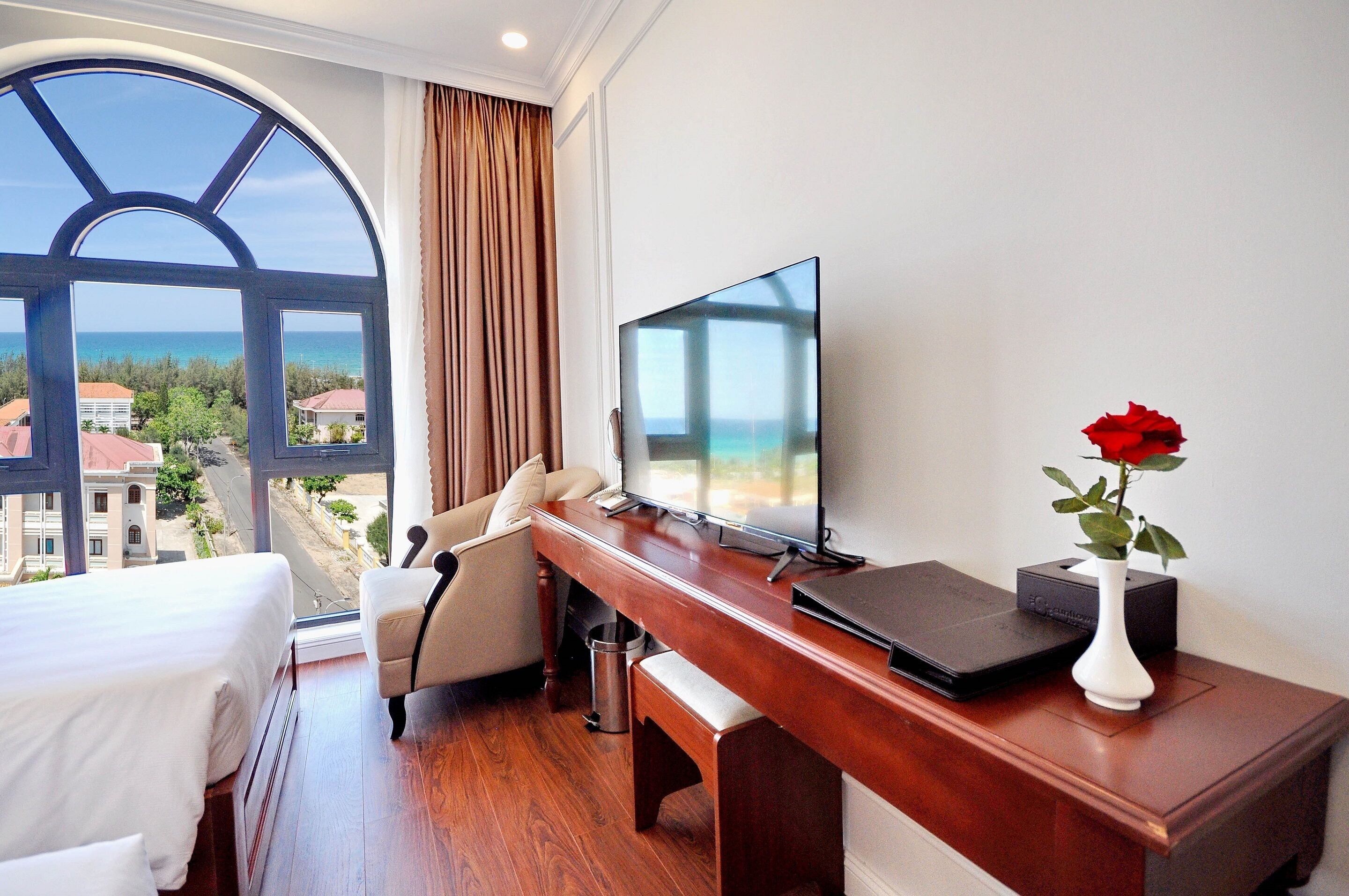Une chambre d’hôtel avec une grande fenêtre, un lit, un bureau avec un ordinateur portable, une télévision et un vase contenant une rose rouge.