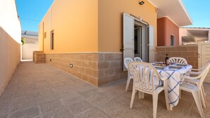 Une terrasse avec une table dressée pour le repas, entourée de chaises blanches, et une porte donnant sur un espace abrité.
