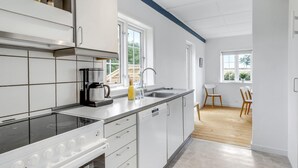 A modern kitchen with white cabinets, a built-in oven, a coffee maker, and a sink. There is a dining area with a table and chairs visible through an open door. - 2 bedroom gorgeous home in Svaneke (Svaneke)