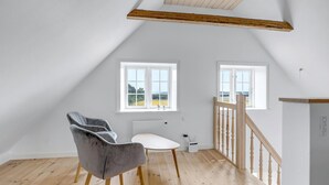 A room with a sloped ceiling, two windows with white frames, a grey chair, a white table, and a wooden staircase railing. - 2 bedroom gorgeous home in Svaneke (Svaneke)