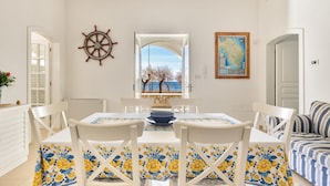 Zona pranzo con tavolo apparecchiato per quattro, sedie bianche, decorazione a timone di nave, una mappa appesa al muro e vista sul mare attraverso un'apertura.