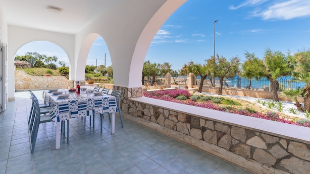 Zona pranzo all'aperto coperta, con parete in pietra e vista su piscina e vegetazione.