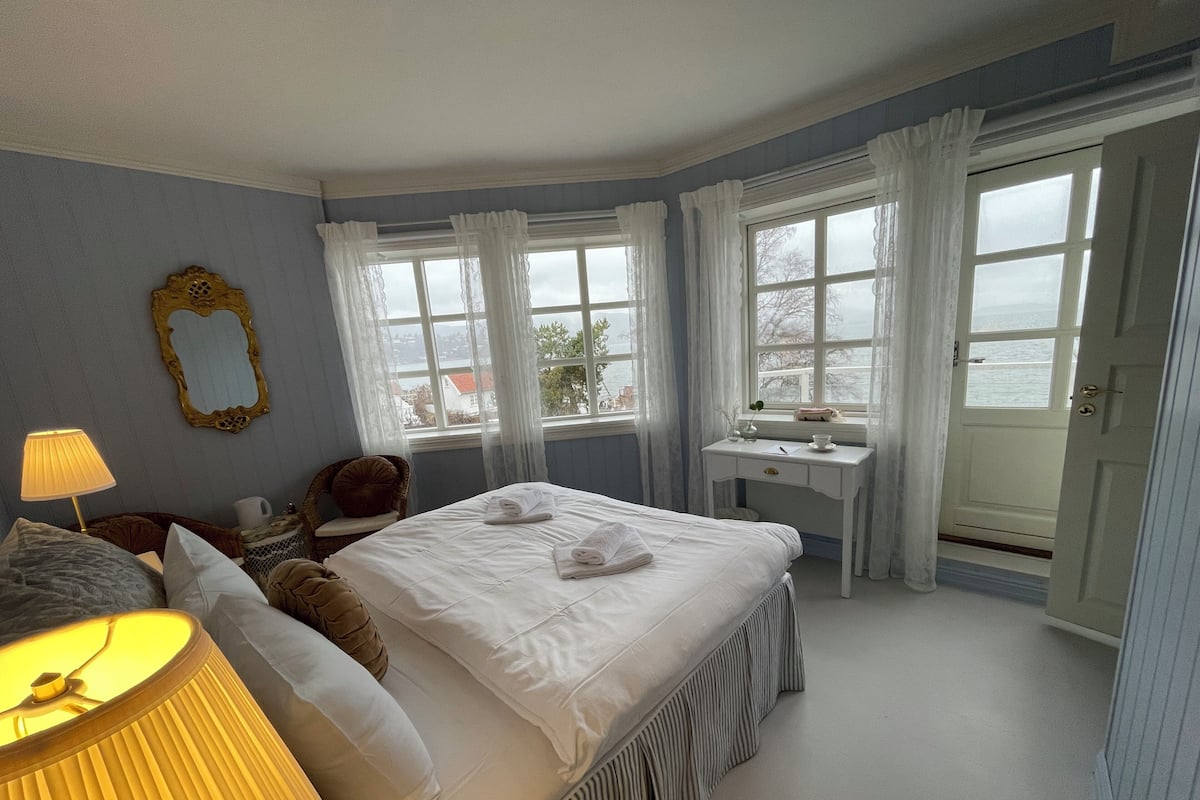 Un dormitorio ordenado con cama, mesita de noche, lámpara y vistas al mar a través de amplios ventanales.