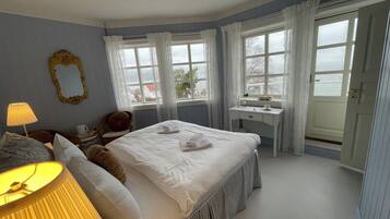 A neatly arranged bedroom with a bed, bedside table, lamp, and a view of the sea through large windows.